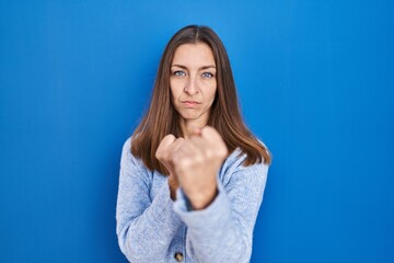 Young woman standing over blue background ready to fight with fist defense gesture, angry and upset...