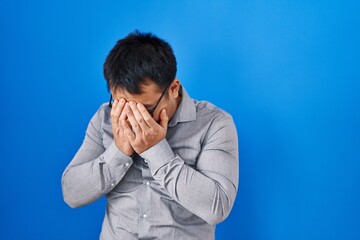 Young chinese man standing over blue background with sad expression covering face with hands while crying. depression concept.