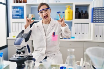 Fototapeta premium Hispanic girl with down syndrome working at scientist laboratory shouting with crazy expression doing rock symbol with hands up. music star. heavy music concept.