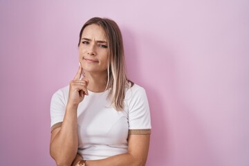 Blonde caucasian woman standing over pink background thinking concentrated about doubt with finger on chin and looking up wondering