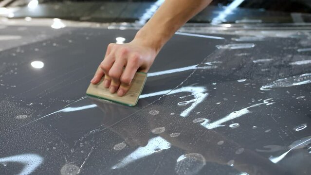 Close up. A female hand with a plastic scraper glues a protective anti-gravel film on the hood of a gray new car. Concept of car body protection with special films.