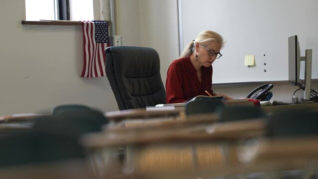 Happy female teacher sitting at desk in empty school classroom desk grading papers working on assignments. US American flag in the background.