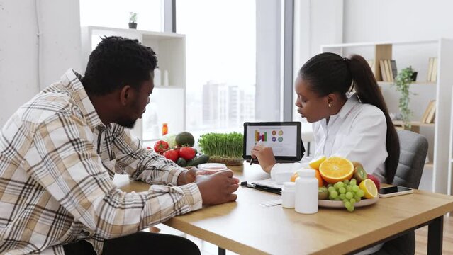 Over-shoulder view of african american woman analyzing patient's nutrition data on digital tablet in consulting room. Efficient dietitian assessing health needs of male client in medical centre.