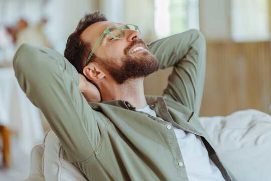 Millennial bearded man, wearing trendy glasses, sitting on sofa and stretching hands