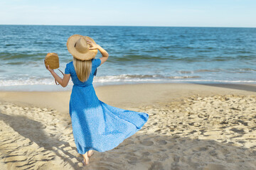 Fototapeta na wymiar Woman in dress with straw hat walking by sea on sunny day, back view