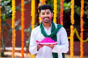 Happy indian man with holi color on face by holding holi color plate by looking at camera - concept...