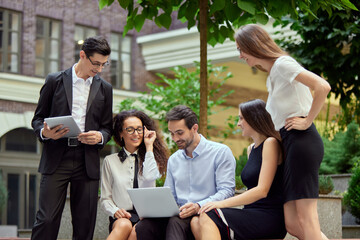 Group of employees, business people, project leaders sitting on bench outside office on daytime and working online on laptop. Business, career development, ambitions, success, office lifestyle concept