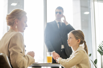 preteen girl talking to blonde mother near bodyguard on blurred background, security service, private safety concept, glass of orange juice, personal safety