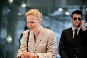 happy blonde woman in formal wear standing at reception desk, personal security service concept,...