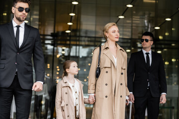 bodyguards walking next to blonde successful woman and preteen kid, entering hotel, private...