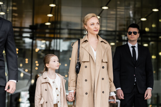 bodyguards walking next to blonde woman and preteen kid, entering hotel, private security, successful mother holding hands with daughter and wearing trench coats, safety and protection