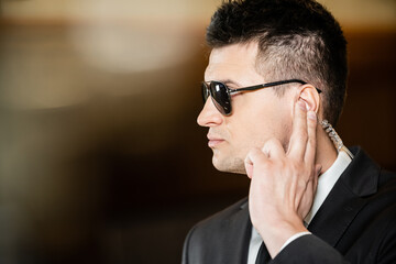 handsome bodyguard in sunglasses, handsome man in suit and tie touching earpiece in lobby of hotel, security, career, communication, vigilance, private safety, hotel work
