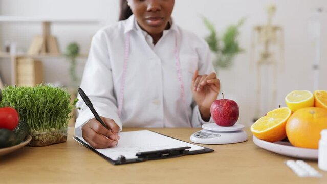 Close up view of health practitioner putting red fruit on digital device for weighing while recording information on paper. Multiethnic nutrition specialist achieving goals with smart technologies.