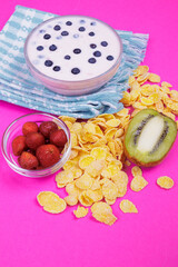 Glass bowl of yogurt with fresh blueberries, bowl with strawberries, half of kiwi and a lot of scattered cornflakes on pink background.