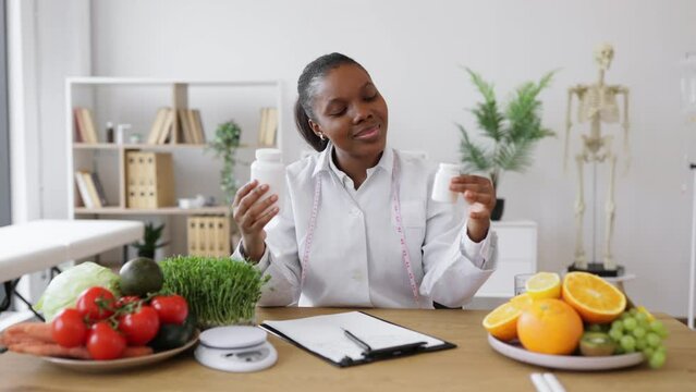 Focused female nutritionist holding two medicine bottles while working at office desk with products on it. Multiethnic health professional choosing food supplement for balancing diet of clients.