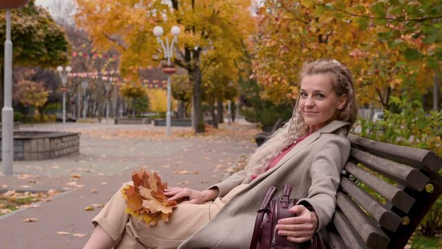 A beautiful mature blonde woman with pigtails in a trendy beige coat sits resting on a bench in a city park. Middle-aged woman enjoying autumn in the city