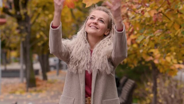 Portrait of a fashionable mature blonde woman with pigtails in a trendy beige coat in a city park in autumn. Happy middle-aged woman enjoys autumn in the city and plays with leaves, throws them up