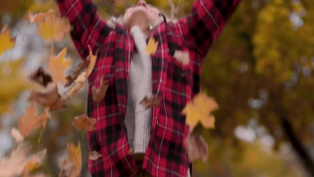 A beautiful mature woman in a red plaid shirt collects leaves in the park in autumn. A middle-aged woman plays with autumn leaves and throws them up.