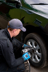 Auto mechanic in a cap changing a wheel from a black car using a drill in a tire shop on the street detailing car repairs