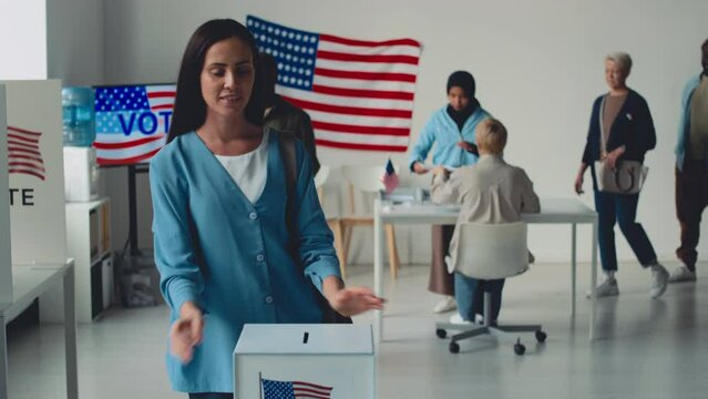 Medium slowmo portrait shot of beautiful young Caucasian woman with long dark hair casting her ballot at electoral precinct in state referendum, looking at camera and smiling, USA emblem in background