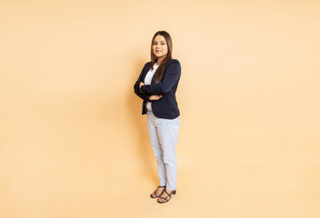 Fototapeta na wymiar Portrait of beautiful young indian business woman wearing formal suit standing with her arms crossed isolated over beige background. Corporate concept. Copy space. Full length.