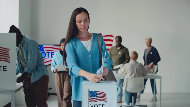 Full shot from ballot box on US state election day, of beautiful young Caucasian woman, African American man, Muslim lady in hijab casting their papers after voting in booth with emblem of USA flagFul