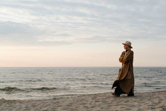 Thoughtful Lonely Stylish Senior Woman In Brown Coat And Hat Walking On Beach In Evening, Copy Space. Adult Caucasian Lady Alone Looking At Sea