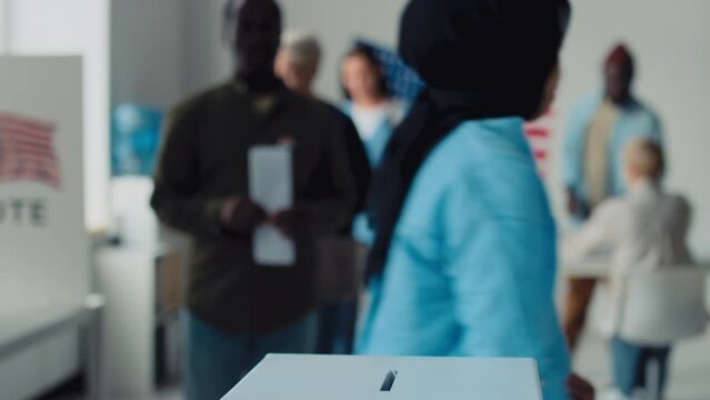 Medium close-up shot of young Muslim woman in black Islamic hijab and blue blouse casting her ballot in box at polling station in USA and leaving, African American man, Caucasian lady queuing behind