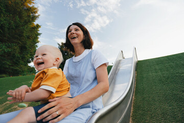 young mother slides down the hill with her toddler son very happy and laughing merrily