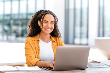 Positive successful female freelancer. Pretty, brazilian or hispanic curly young woman, university student, sitting near campus, with laptop and notebook, preparing for exam, looks at camera, smile