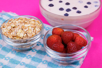 Glass bowl of yogurt with fresh blueberries bowl with strawberries and bowl with cereals on pink background.