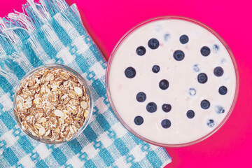 Glass bowl of yogurt with fresh blueberries and bowl with cereals on pink background. Top view.