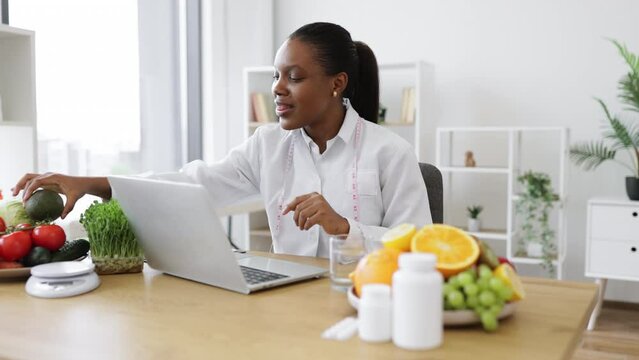 Charming adult lady in lab coat showing avocado to front camera of computer in professional office of clinic. Multicultural specialist in nutrition counseling patient online about benefits of fruit.