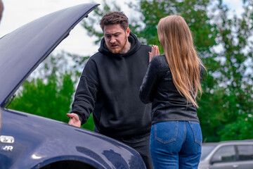 Fototapeta na wymiar A young couple stands near a broken car with an open hood A man explains what has broken in a parking lot against the background of trees