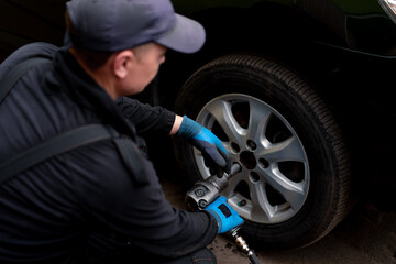 Auto mechanic in a cap changing a wheel from a black car using a drill in a tire shop on the street...