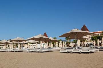 Row of sun loungers and umbrellas on the empty beach during sunrise awaits tourists. Blue sky background