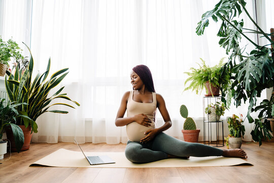 Black pregnant woman smiling and using laptop during yoga practice