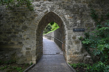 Fototapeta na wymiar Stone wall with a pointed arch passage. On the background there is a path and railings. The path leads to wood. On the stone wall there is a shield with the street name, it is called s' Hintertörli. 