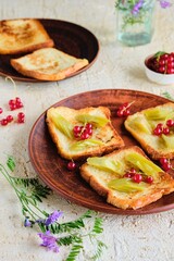 Dessert or breakfast, fried toast with rhubarb in sugar syrup and fresh red currants on a brown plate on a beige concrete background. Seasonal recipes, rhubarb dishes.