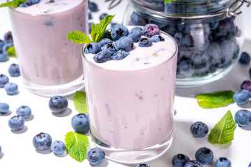 Blueberry yogurt, smoothie or milkshake on portioned glasses with fresh blueberries decor on it, and a lot of fresh berries on white kitchen table background copy space