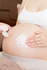 a pregnant girl sits at home on the bed and smears an anti-stretch mark cream on her stomach. Pregnancy, motherhood, preparation and expectation concept