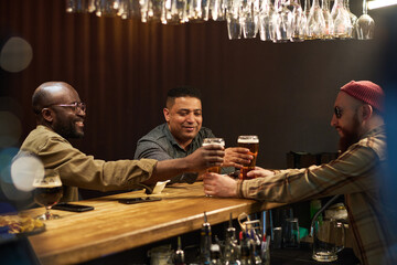 Two happy intercultural men taking glasses of fresh beer from hands of bearded barman in sunglasses standing by counter in front of them