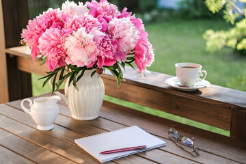 A vase with peonies, a notebook and a cup of coffee on the terrace in the summer garden.