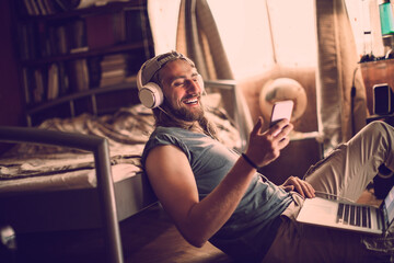 Young man using a smart phone and laptop whie listening to music in the bedroom