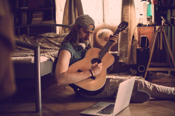 Young man playing his guitar while using a laptop in his bedroom
