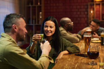 Happy young brunette woman with glass of aperitif toasting with her boyfriend while both sitting by...