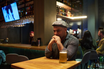 Young depressed man in casualwear keeping hands by his face while sitting by table with big mug of beer against bar counter and other people