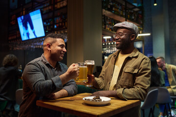 Two happy intercultural buddies clinking with glass and mug of foaming beer while sitting by table in the bar and enjoying weekend