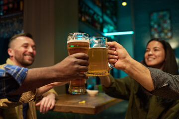 Focus on hands of intercultural buddies clinking with glasses and mugs of refreshing beer while sitting by table and bar counter and toasting