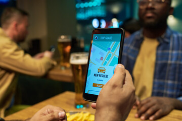 Hand of young man holding mobile phone while sitting in front of his buddy in pub, choosing and ordering taxi with help of application